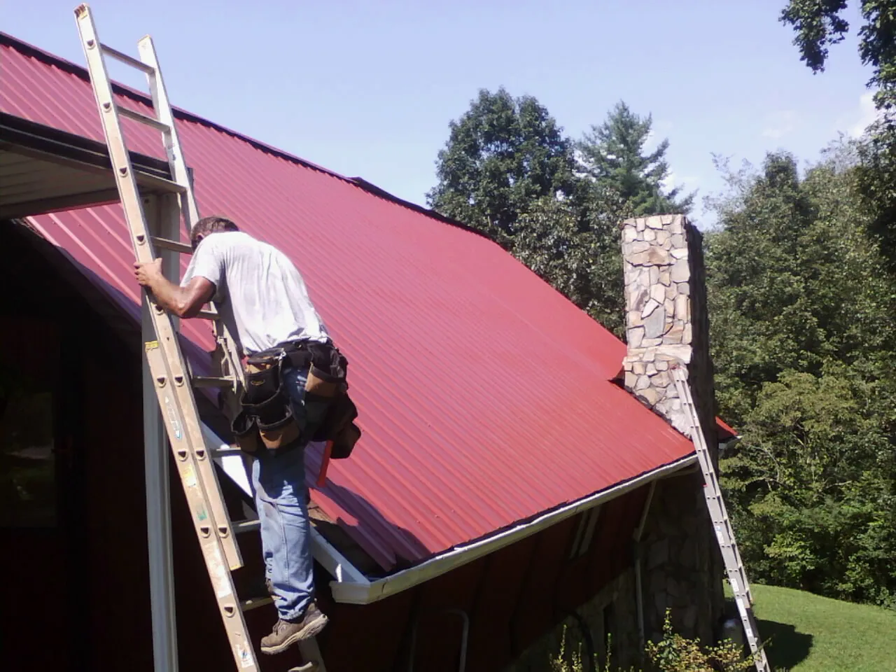 Roofer on a ladder beside a red metal roof and stone chimney — Soffit & Fascia Repair by Roofing Brothers in Roby